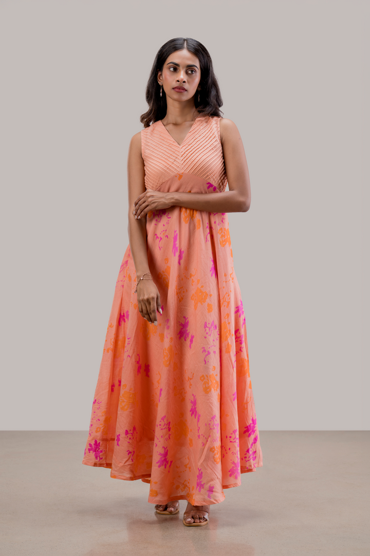A woman modeling a sleeveless peach maxi dress with a pleated bodice and a flowing skirt featuring orange and magenta abstract floral patterns, set against a neutral studio backdrop.