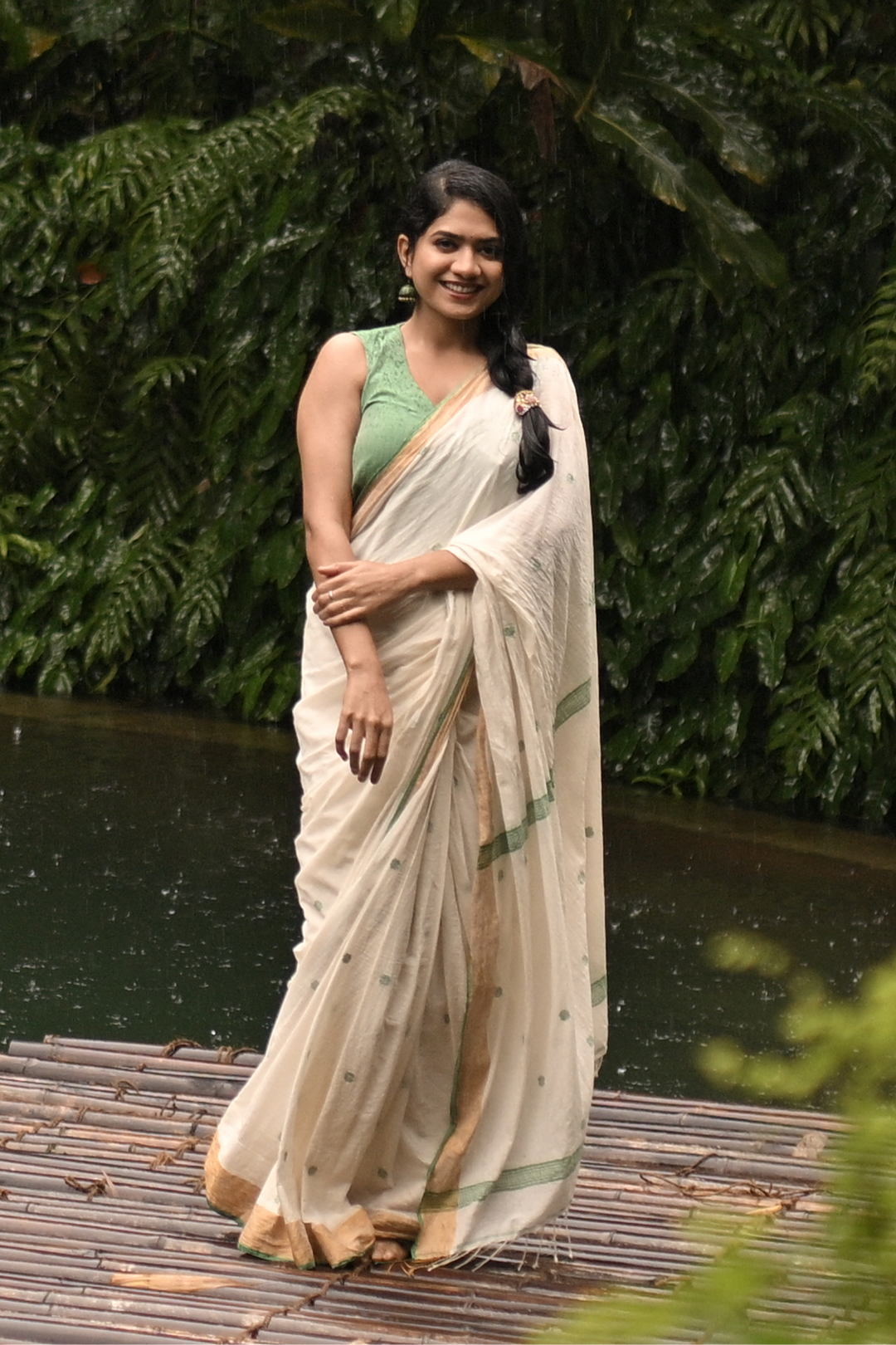A woman wearing a light beige and green saree stands on a bamboo raft over calm water, with dense green tropical foliage in the background.