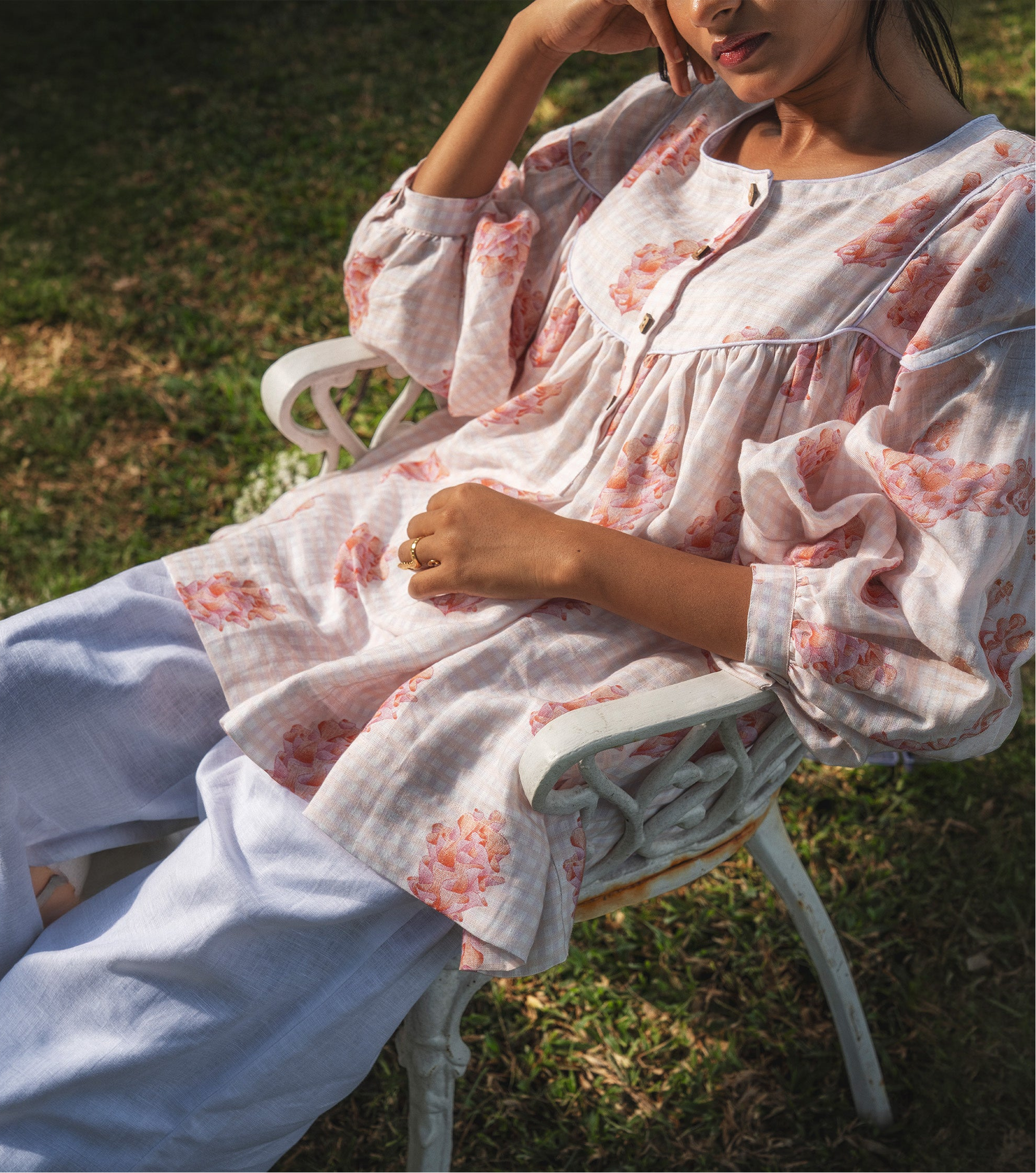 Person sitting outdoors on a chair with a floral dress and light blue pants.
