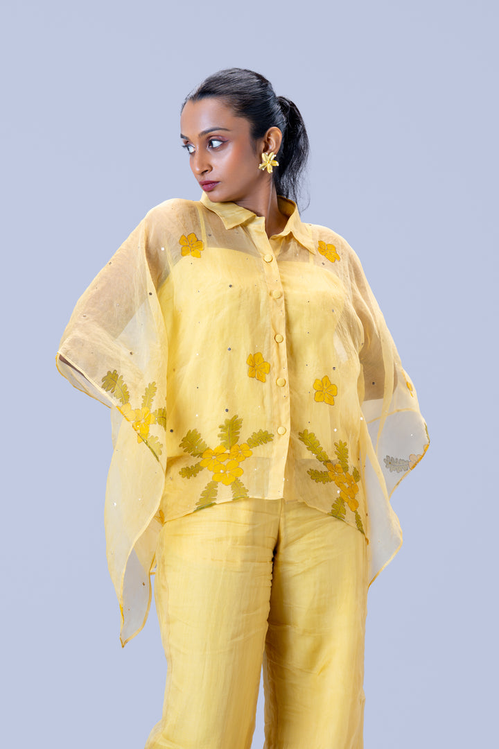 A woman modeling a light yellow two-piece outfit with a matching sheer embroidered dupatta, featuring yellow floral patterns, set against a solid light blue-gray background.