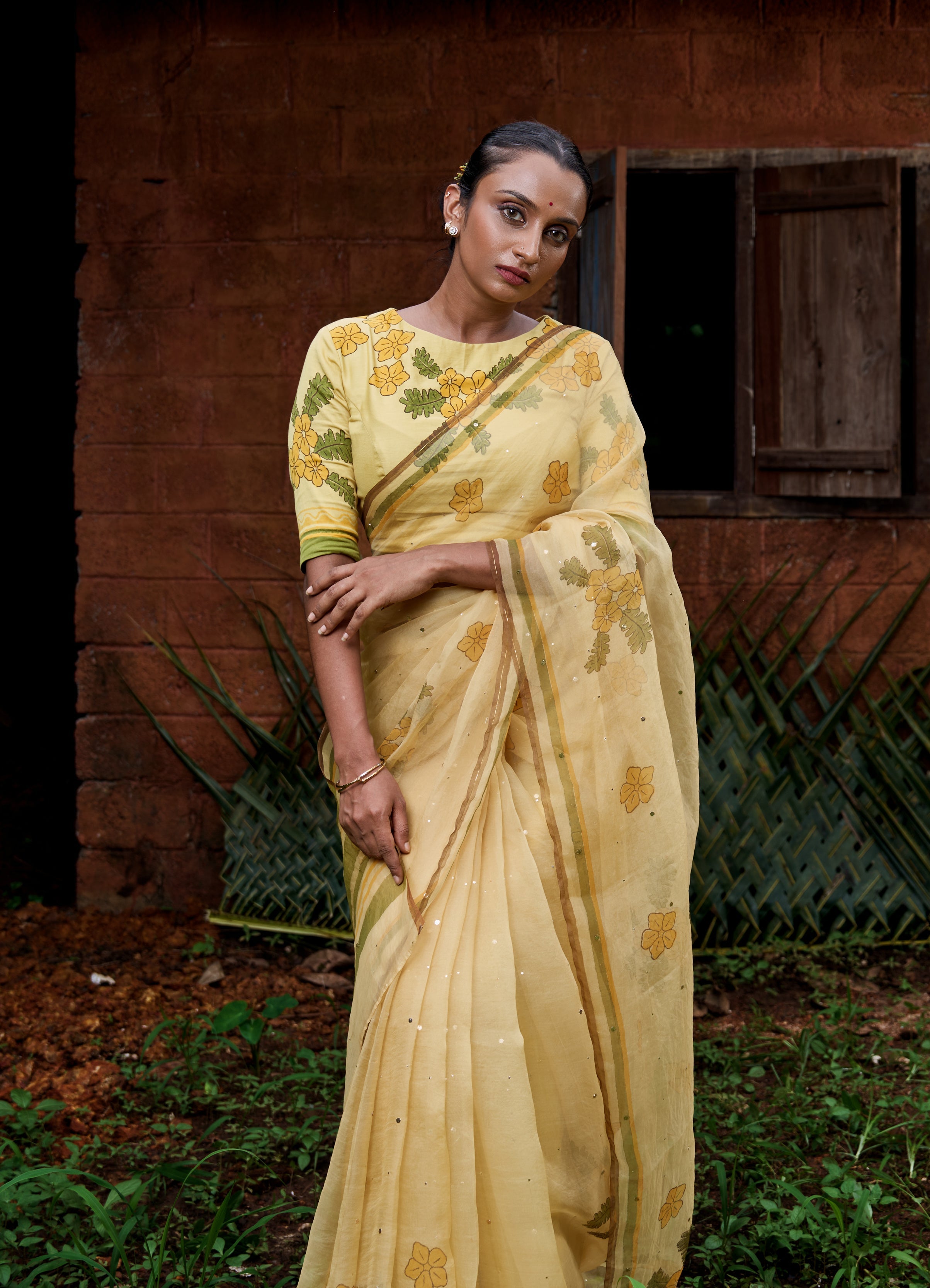 Two women wearing yellow sarees with green floral patterns pose outdoors in front of a rustic red-brown brick wall with wooden shutters.