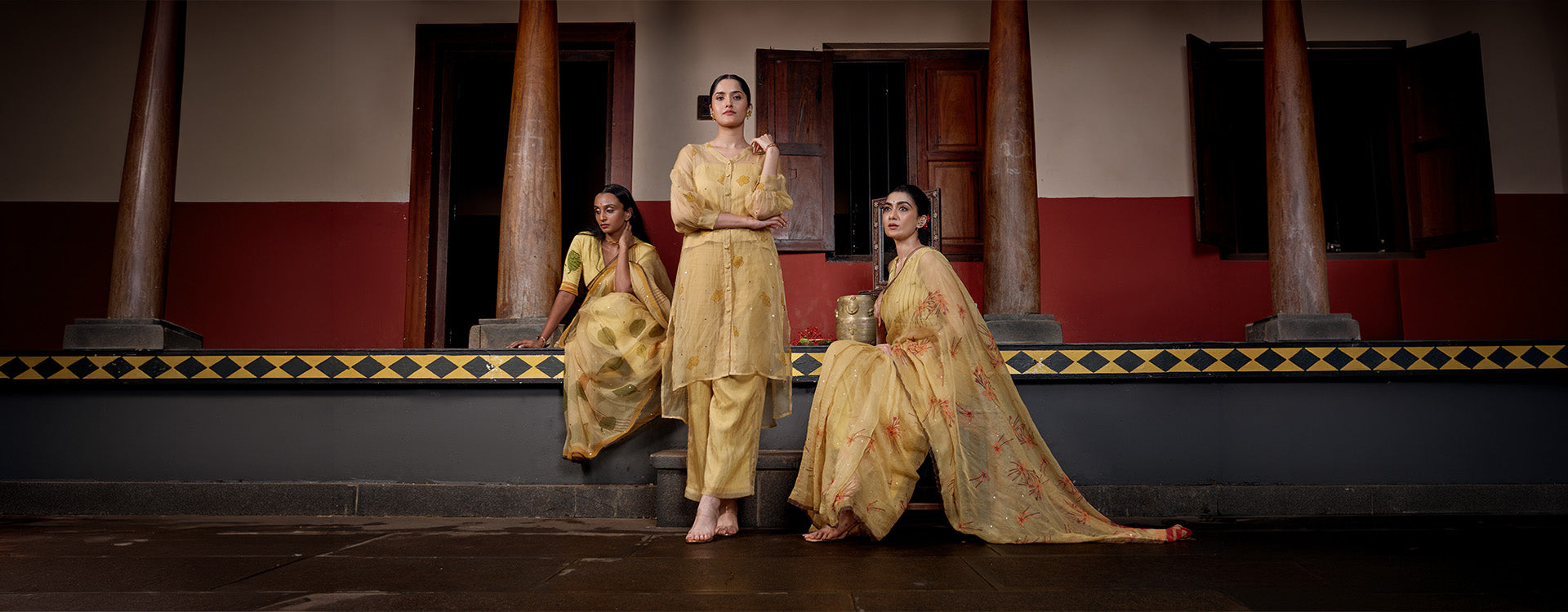 Three women modeling traditional Indian salwar kameez in a matching golden-yellow fabric, posed on steps in front of a red and brown architectural backdrop.