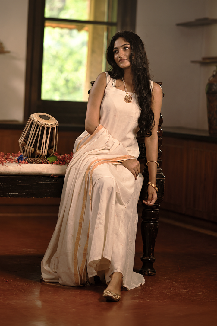 A woman wearing a white sleeveless dress and a white saree with gold border details sits on a dark wooden chair in a traditional Indian room. She is adorned with gold jewelry, and the background features a wooden bench with traditional drums and decorative vases.